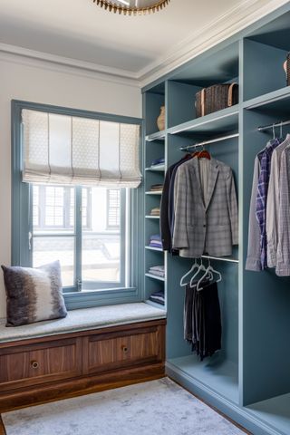 walk-in dressing room with blue storage shelves and window seat with shirts and trousers hanging