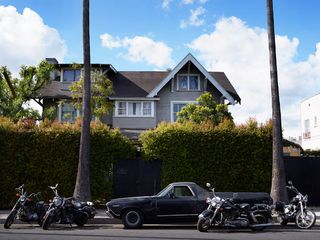 Daytime image, parked black car and four retro style motorbikes, trunks of two palm trees, tall evergreen hedges with solid black gates in the centre and to the right, top of detached house, with windows and roof protruding over the hedge, tops of trees, cloudy blue sky