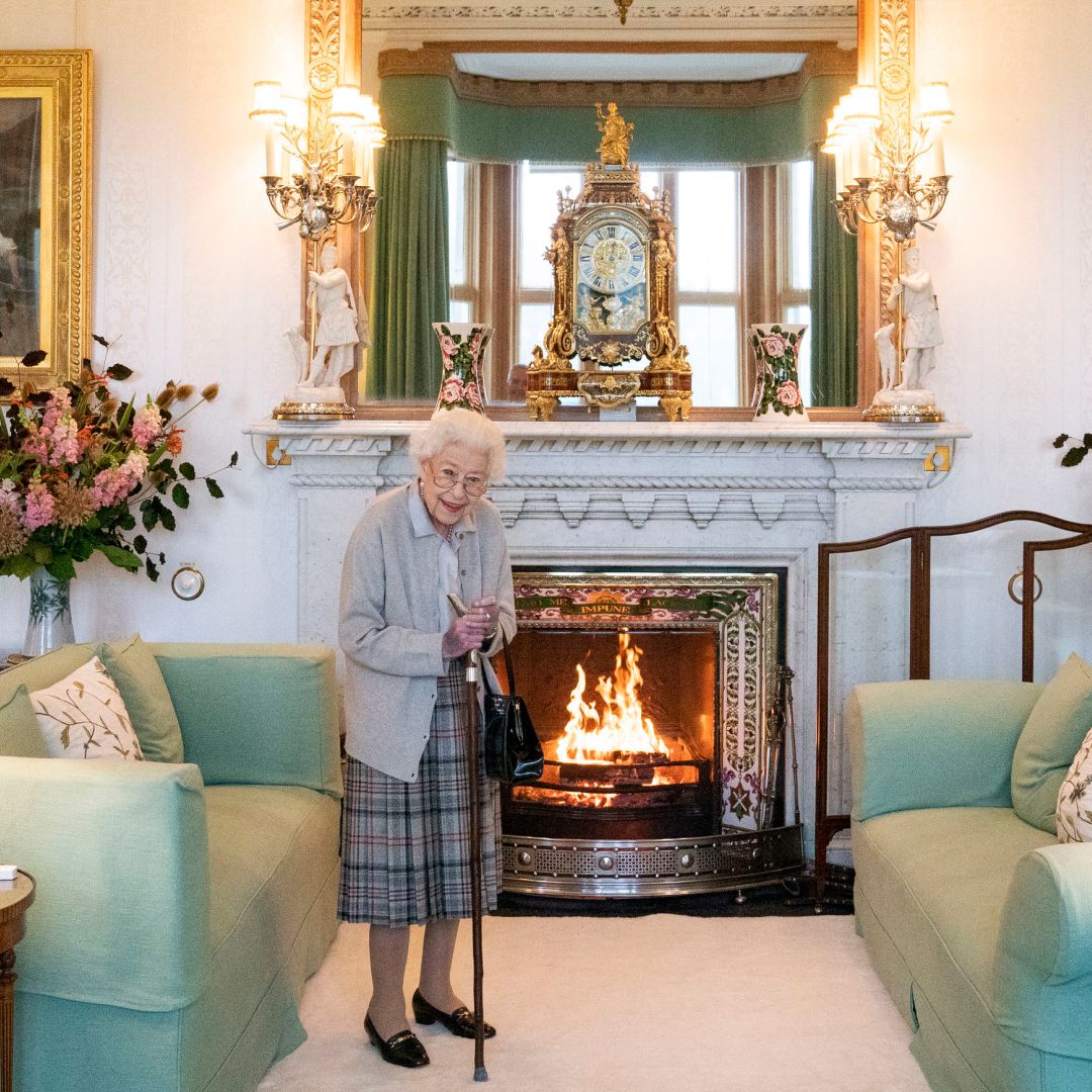 Queen Elizabeth wearing a plaid skirt standing in front of a fireplace and green sofa