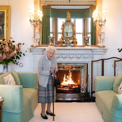 Queen Elizabeth wearing a plaid skirt standing in front of a fireplace and green sofa
