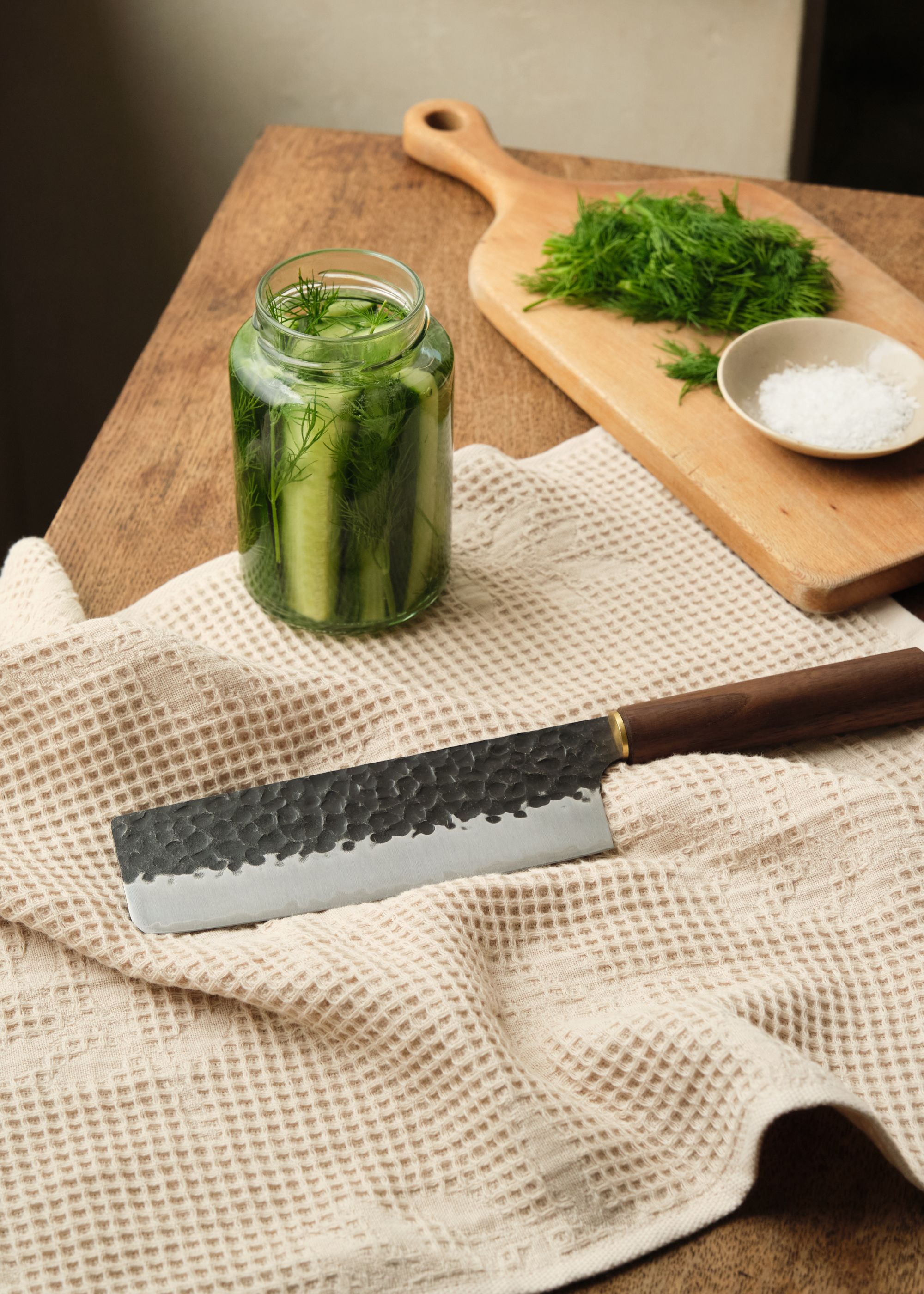 A Japanese knife on a cloth by a jar of cucumbers and dill and a cutting board with dill and a bowl of salt