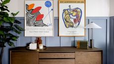 Entryway with blue wood panelled wall and wood console with lamp and books and two vintage posters