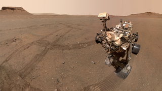 photo of a large white and silver mars rover sitting on the red dirt, with several small buttes in the background