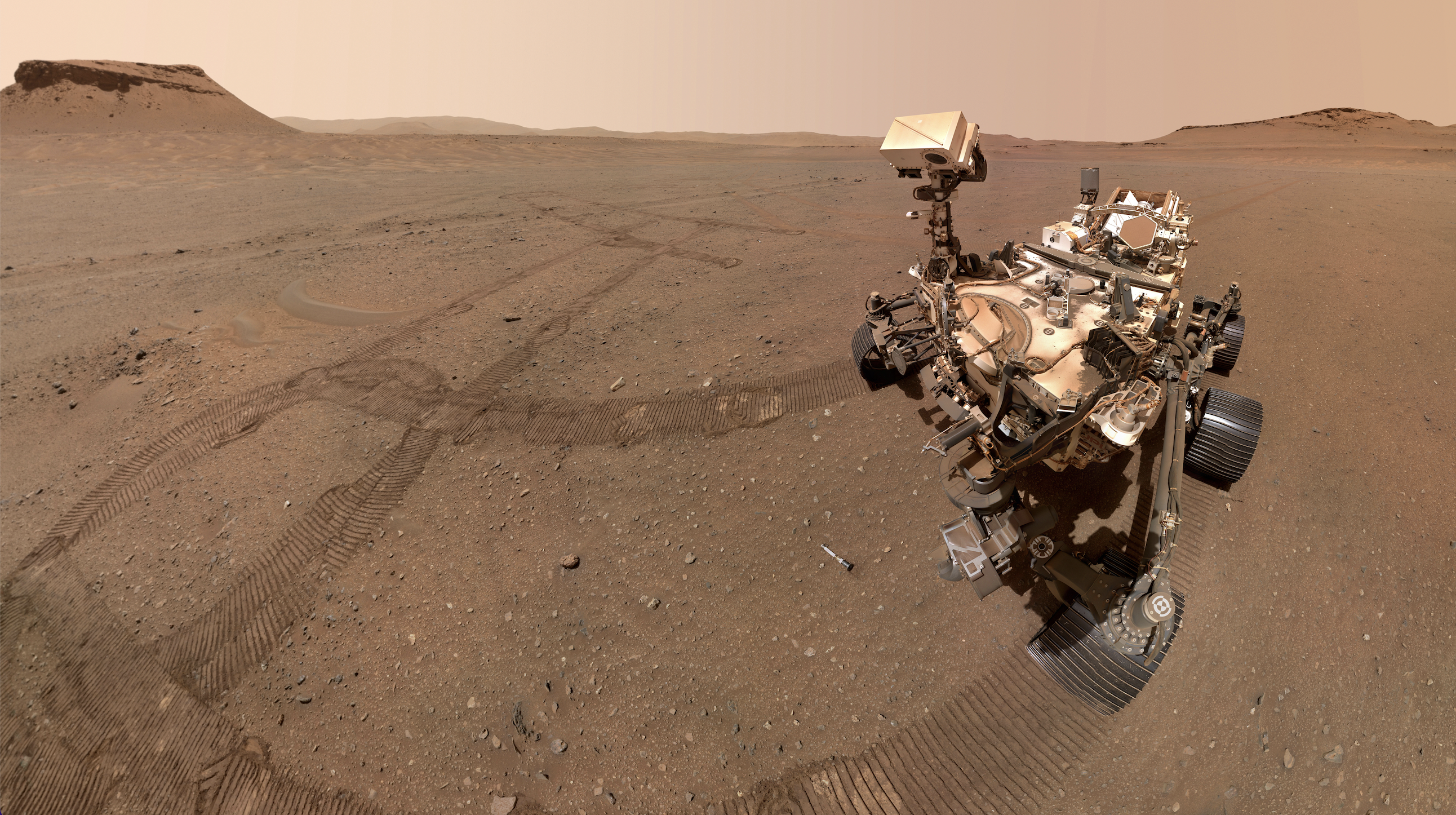 a large mars rover sits in a sandy patch of red planet ground, with some of its tire tracks nearby.