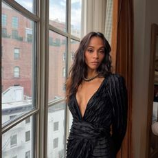 Zoe Saldana wears a black dress and stands in front of a window