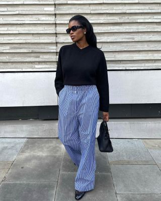 British influencer Marilyn Nwawulor-Kazemaks poses on a London sidewalk wearing black sunglasses, a black sweater, blue striped pajama pants, and black Chanel ballet flats.