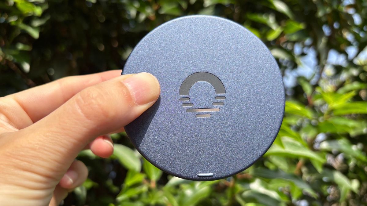 A hand holds the battleship blue Ozo Sleepbuds 2 case against a backdrop of green foliage