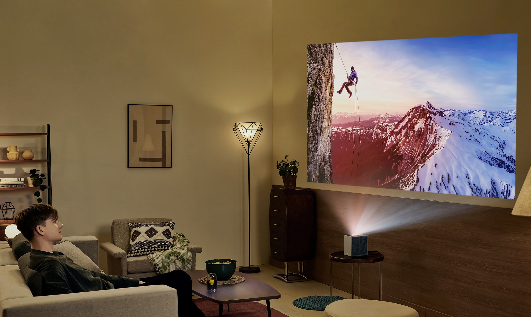 A white living room, with a young white male sat down on the sofa in the far left corner. In front of him, the LG CineBeam S projects an image of a climber abseiling down a snow-tipped mountain.