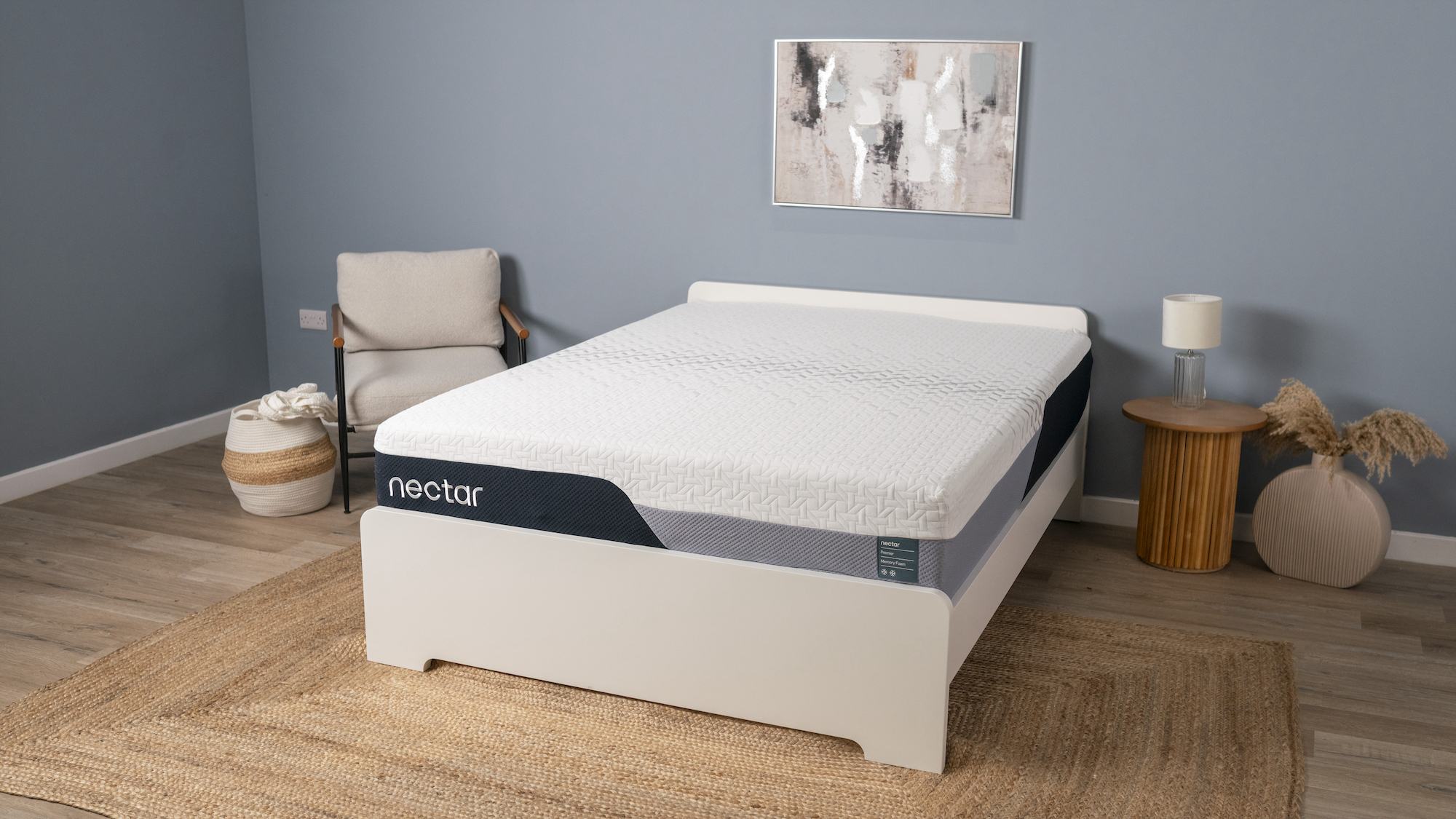 The Nectar Premier Memory Foam Mattress photographed on a white wooden bed frame in our blue Sleep Lab, with a stylish beige fabric armchair next to the bed and a large rattan mat underneath it