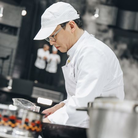 a chef in a white baseball cap and white coat cooks over a silver counter in a still from culinary class wars season 2