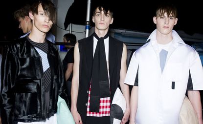 Guys wearing Man S/S 2015 collection. The guy on the left is wearing an apron-like blue and black shirt and a black jacket over it. The middle guy is wearing a white top with red and white blocks at the bottom with a black scarf and a black sleeveless overcoat. The guy on the right is wearing a white top with a blue image on it and a white jacket with some black in it.