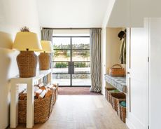 Large entryway with glass and black metal doors, white gloss console table with two large matching lamps, wooden floor, curtains beside door, bench with cushion, storage baskets