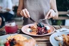 Woman sitting at dining table eating pancakes with blueberries and whipped cream in cafe