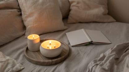 Two bowl candles on a wooden board on a bed with a book and pillows
