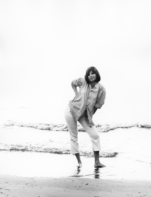 A black and white photograph of Barbra Streisand posing on a beach in 1963, wearing a striped button-down shirt and light-colored trousers.