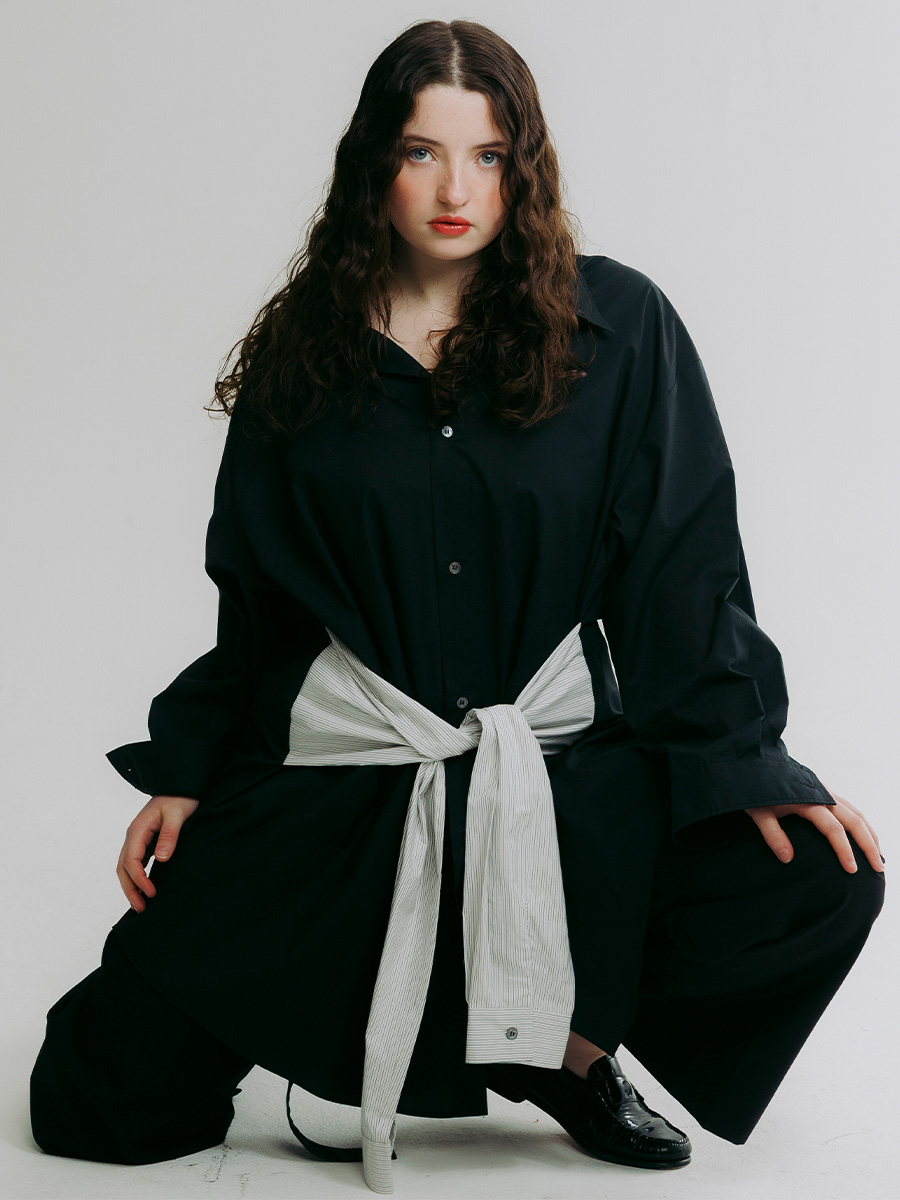 Lucy Halliday photographed by Phill Taylor. She is wearing an oversize black button-down shirt and black trousers with another striped shirt tied around her waist.