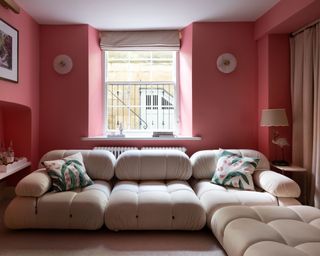 Pink painted living room with large cream sofa with a quilted design, two frosted glass, rounded wall lights and table lamp with scalloped shade, cream blind and pink, green and white cushions on sofa