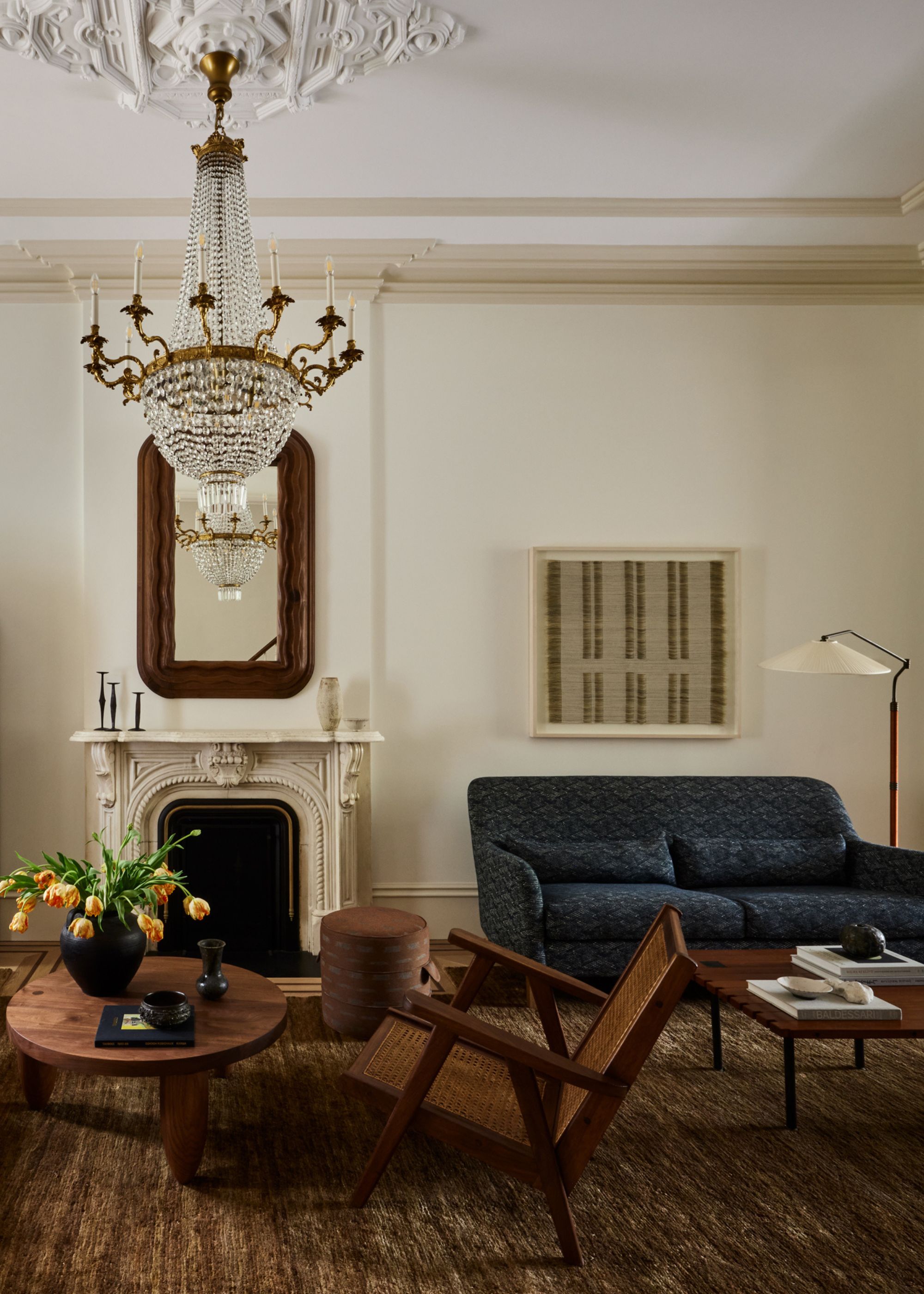 A living room with a chandelier, a framed mirror, a stone fireplace, a vase of tulips, a brown area rug, a rattan armchair, a dark blue sofa, and a floor lamp