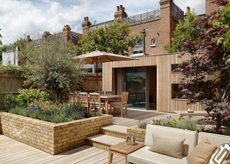 an urban garden with wooden decking, wild flowers and stylish outdoor furniture. In the back of a garden is a wooden garden room with large glass windows