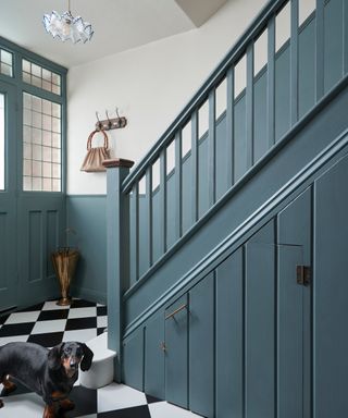 hallway with blue painted door, wall panelling and stairs, black and white tiled floor, white ceiling light and black dachshund dog stood by stairs