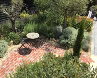 Planting factored in when designing a patio, with olive trees and bushy herbs around a brick patio with bistro table and chairs.