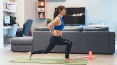 woman doing a forward lunge wearing black leggings and blue crop top on a green mat with a living room setting behind her