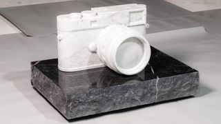 A sculpture of a classic Leica-style rangefinder camera, carved entirely from white marble, rests on a rectangular plinth of rough-cut, dark grey or black marble.