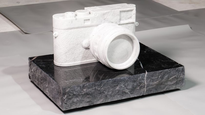 A sculpture of a classic Leica-style rangefinder camera, carved entirely from white marble, rests on a rectangular plinth of rough-cut, dark grey or black marble.