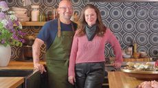 Chef Michael Symon and his wife in front of a navy blue and white tiled kitchen backsplash, showcasing Symon's new mango wood and brass tableware collection
