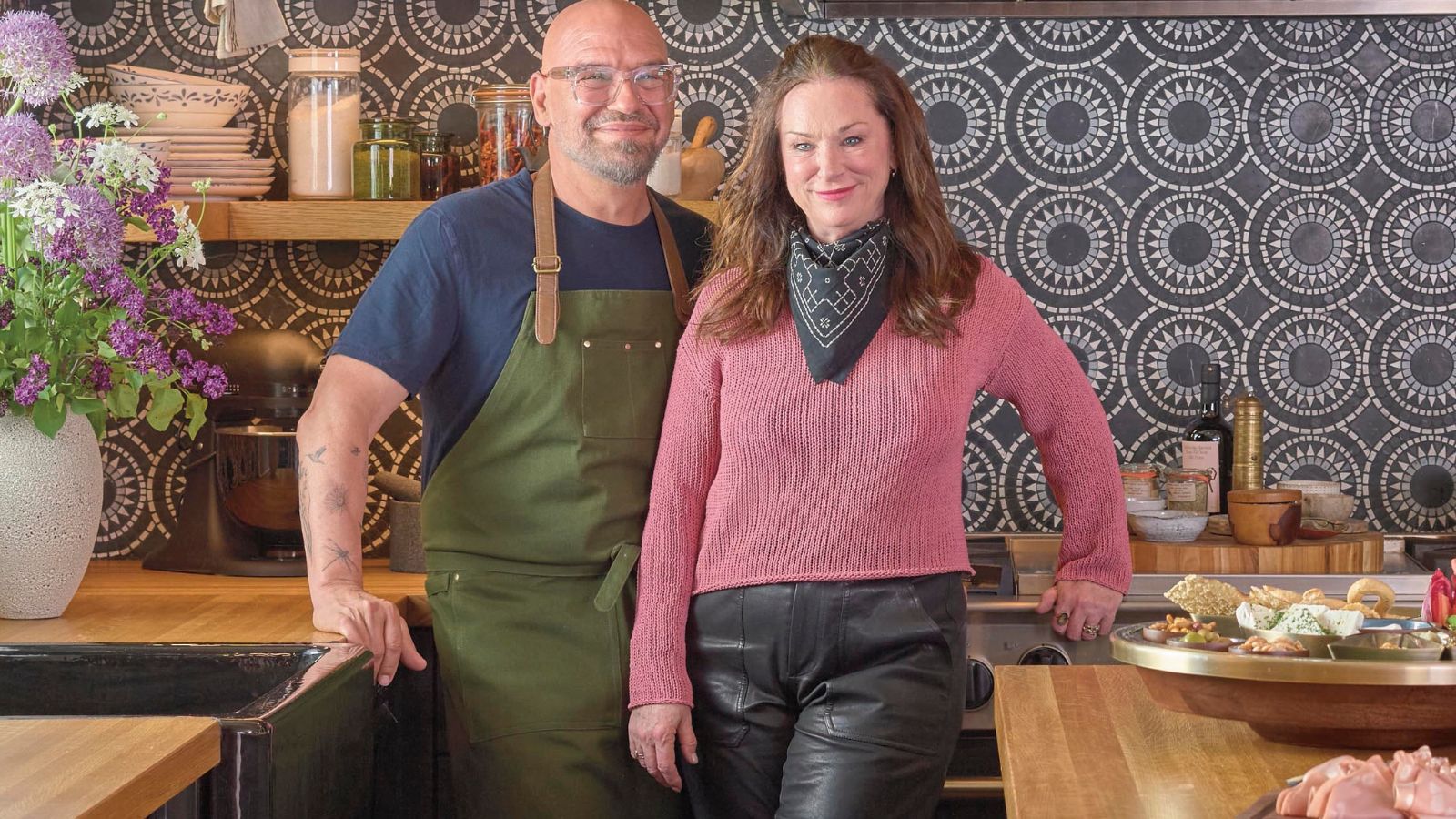 Chef Michael Symon and his wife in front of a navy blue and white tiled kitchen backsplash, showcasing Symon&#039;s new mango wood and brass tableware collection