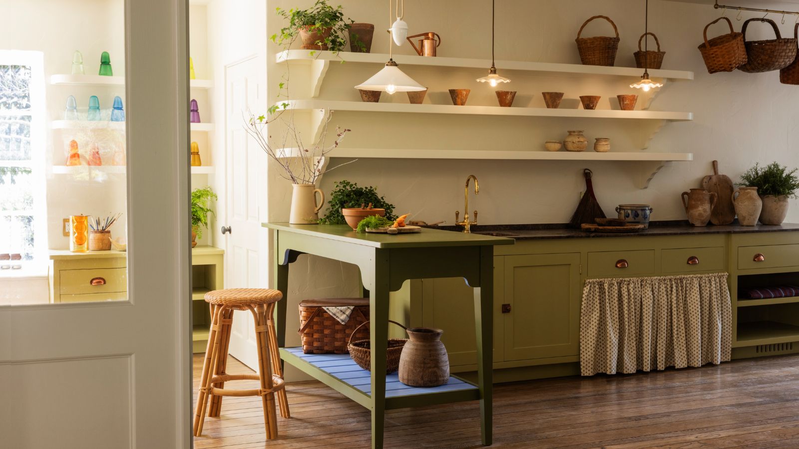 Furniture-led kitchen with a freestanding green island table, open shelving, skirted base cabinets and woven baskets, creating a relaxed, traditional feel.