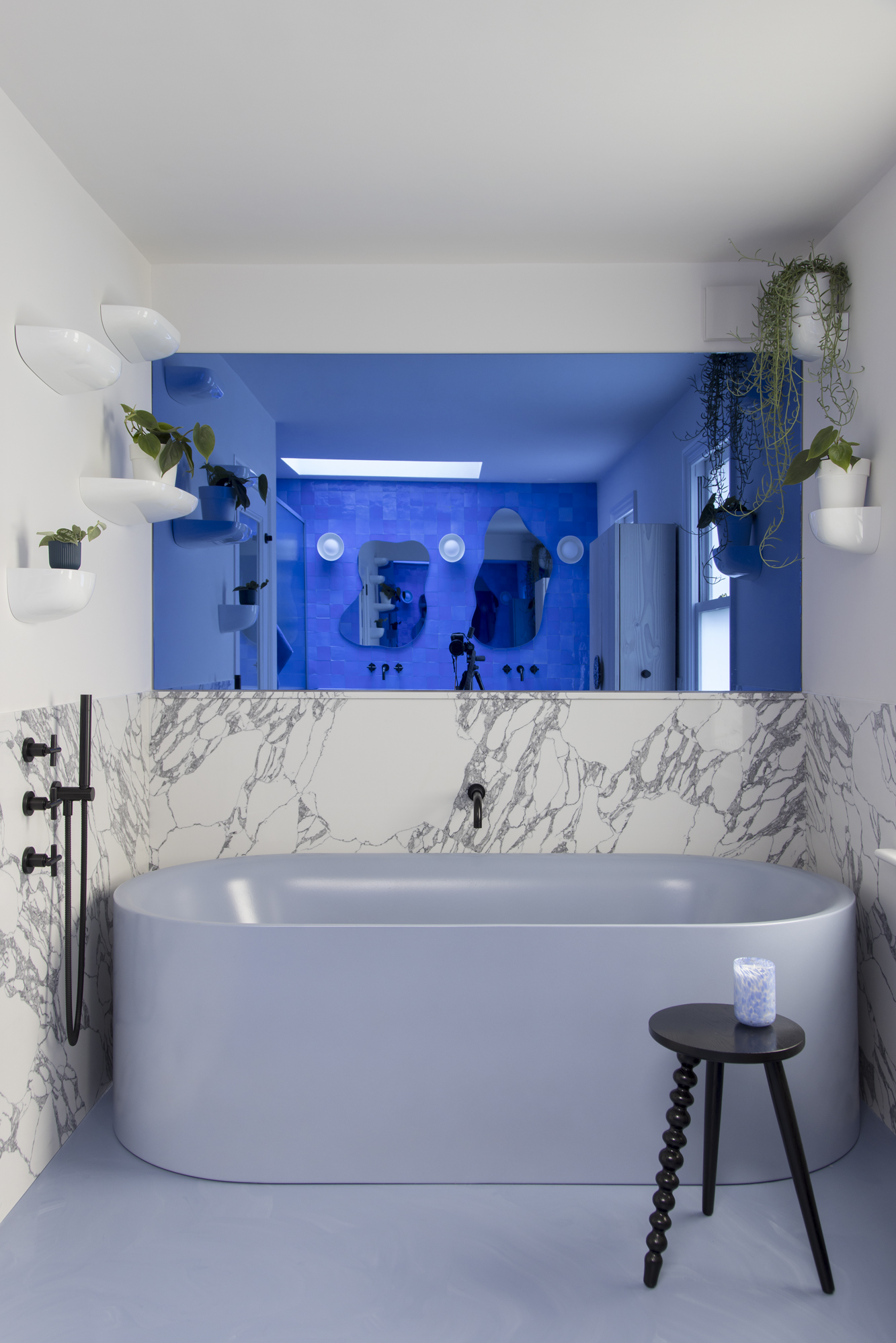 blue bathroom with blue mirrored walls, marble wall, blue freestanding bath, blue floors with black stool next to the bath, floating shelves with plants
