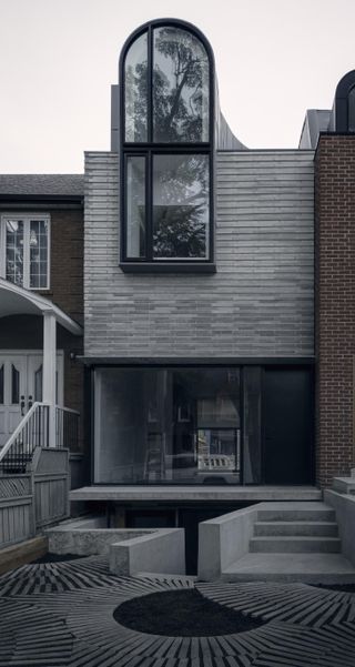 Close-up of a porch and grey courtyard with steps leading up to a two / three story building with grey bricks, patio windows and an oval window at the upper floor.