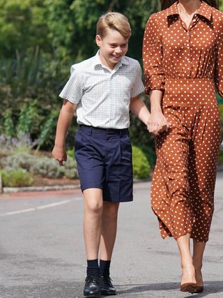 Prince George wearing a white shirt, navy shorts and navy socks with dress shoes smiling and holding hands with Kate Middleton, who is seen from the neck down in a brown polka dot dress walking on a sunny day