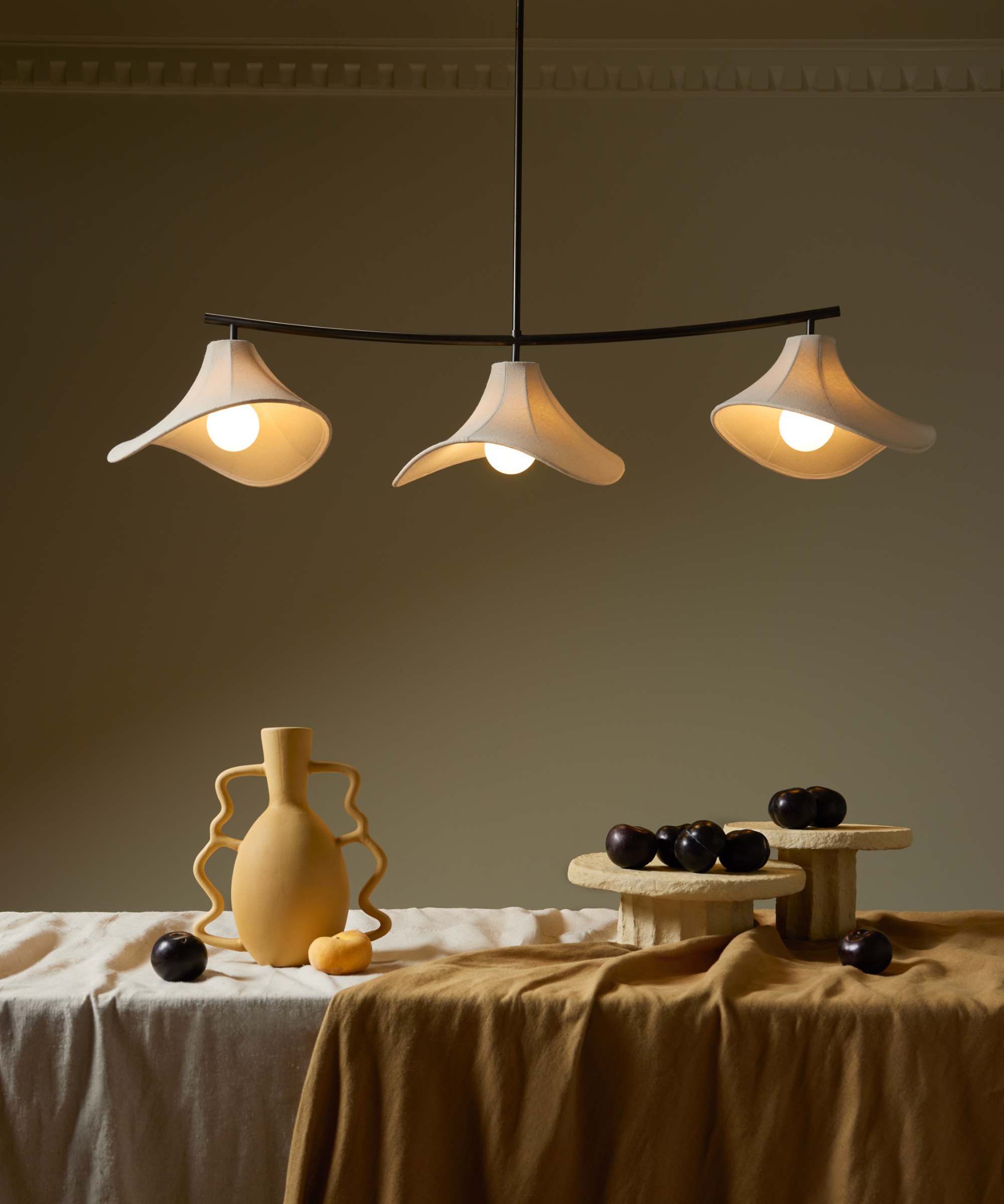 Eny Lee Parker x Lulu and Georgia linear chandelier glowing overhead a long, undone tablescape styled with sculptural objects and scattered fruit.