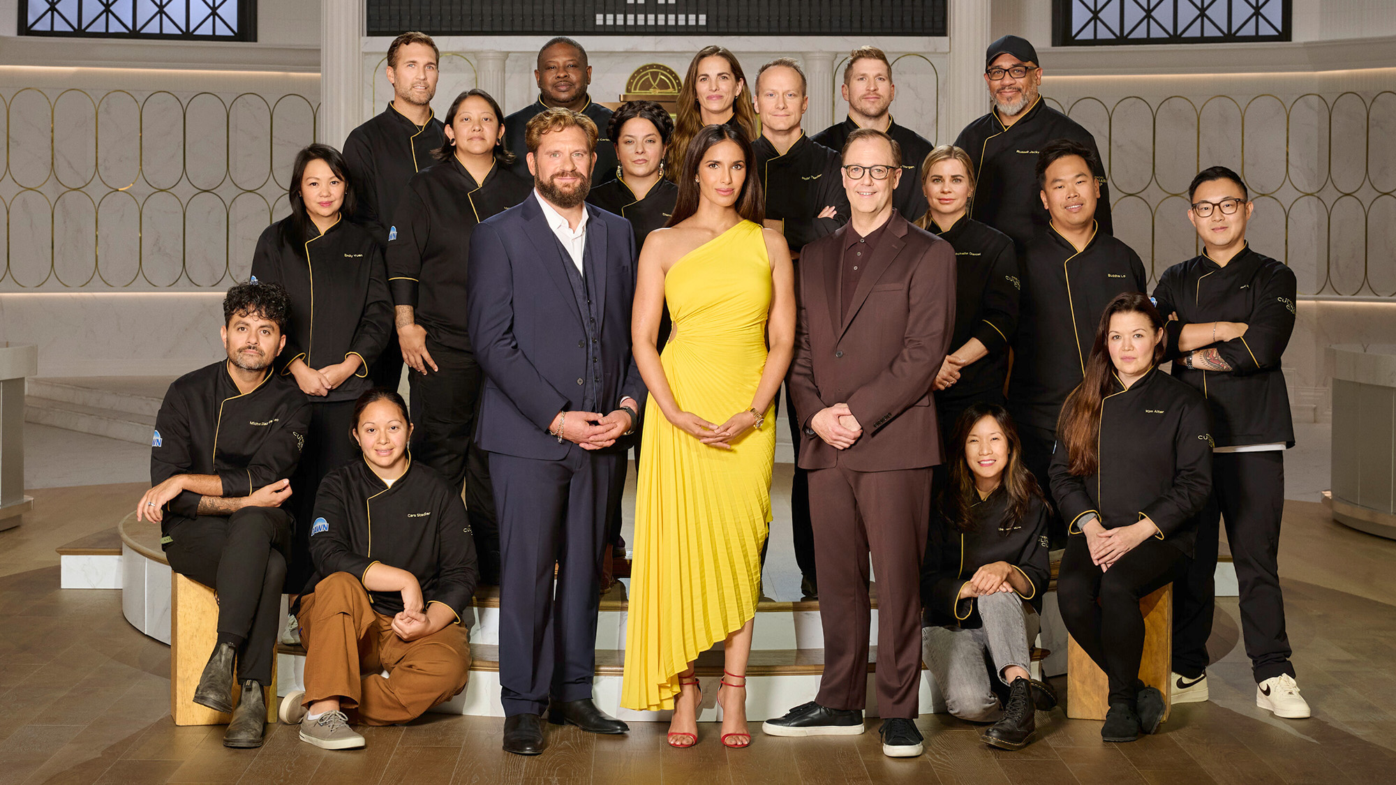 Padma stands in a yellow dress in front of the cheftestants with Michael and Wylie in suits in ACC Season 1.