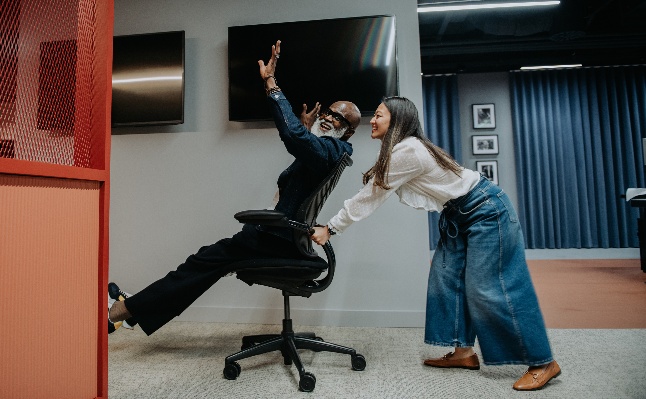 Lighthearted and comical image of two good humored colleagues getting up to some mischief in the office. One pushes the other up the office hallway on an wheelie office chair as the other one enjoys the ride.