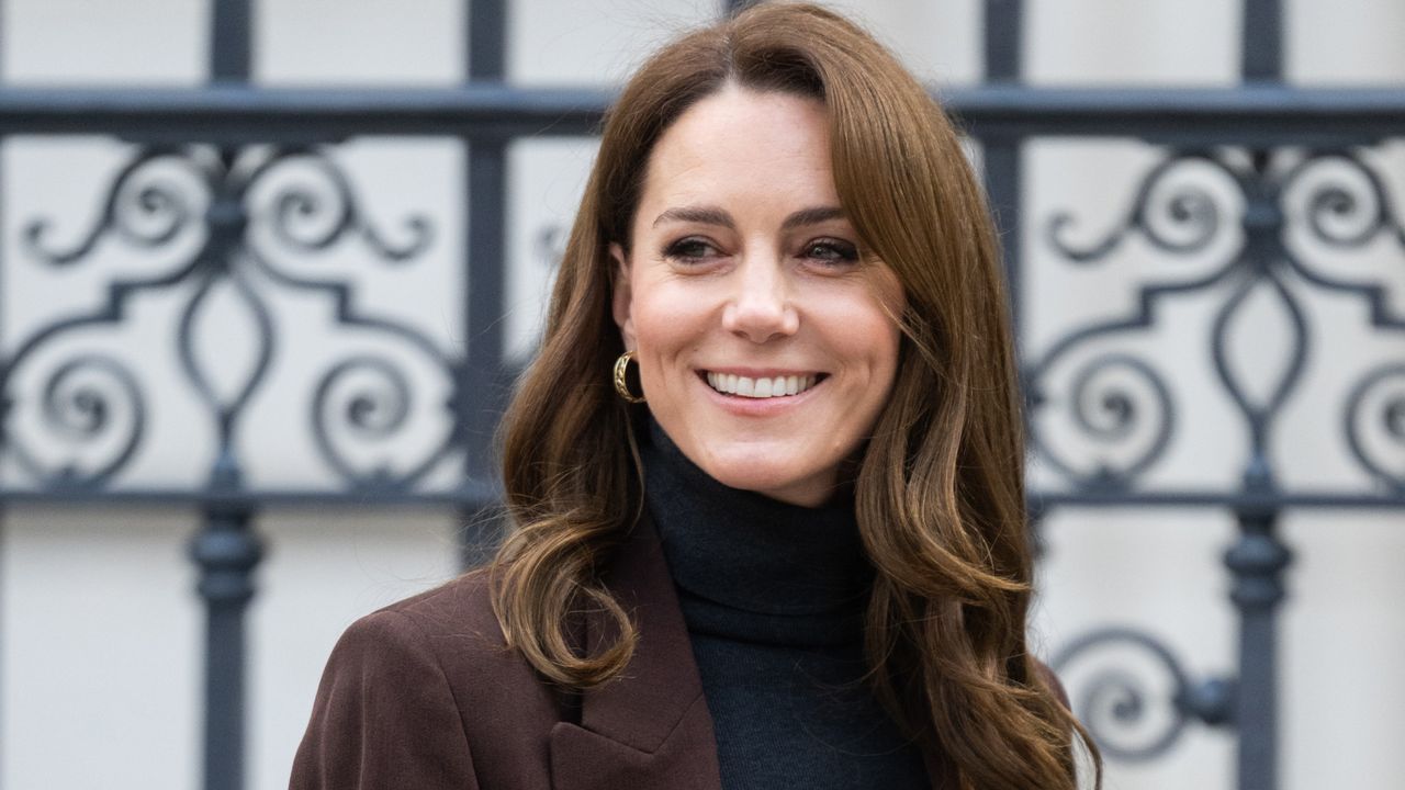 Kate Middleton wearing a brown blazer and black turtleneck smiling and looking to the left in front of a metal gate