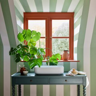 A room decorated in a green and white striped wallpaper with a window frame painted in bright orange and a console with a sink on