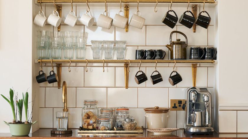 A styled coffee station featuring brass wall shelves with hanging mugs, glassware, storage jars and a stainless steel coffee machine against white tiled walls