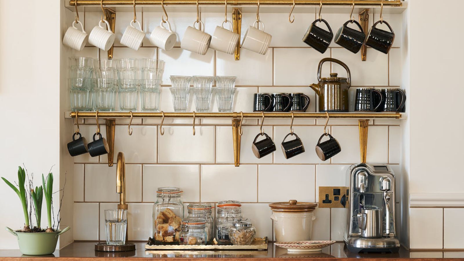 A styled coffee station featuring brass wall shelves with hanging mugs, glassware, storage jars and a stainless steel coffee machine against white tiled walls