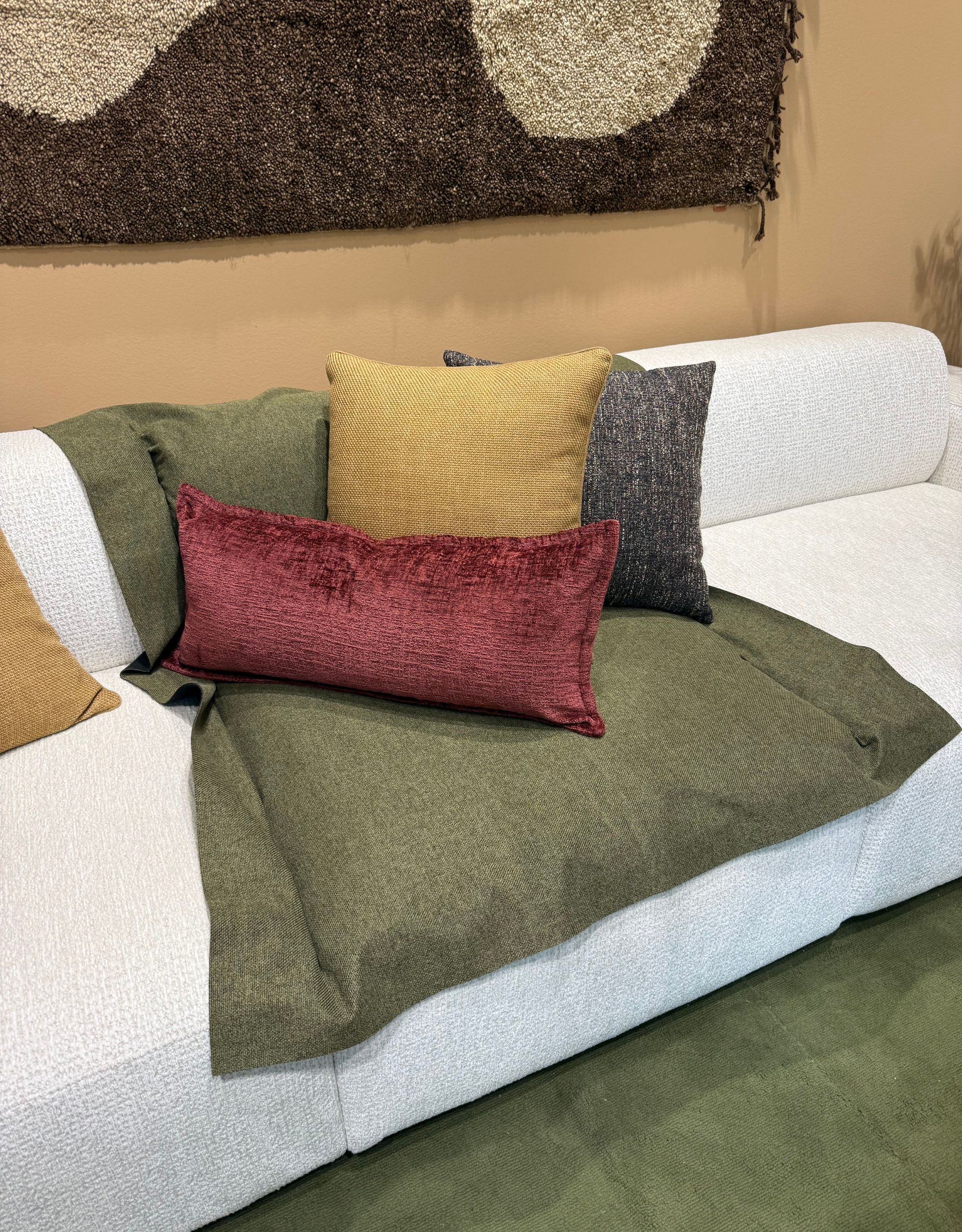 A white sofa with a green fabric placed over the middle and three cushions in different colors resting on top with a rug as art on the wall