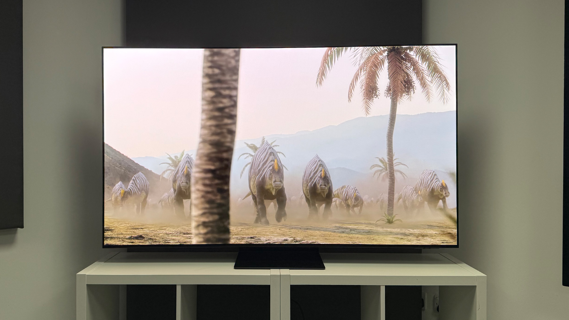 Samsung S95F OLED TV on white wooden furniture, on screen are stripey dinosaurs and palm trees