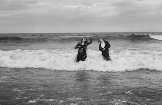 A black and white photograph of two women standing in the sea and laughing as a wave crashes into them