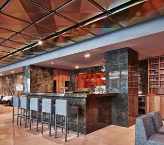 Restaurant with dark grey marble bar, high chairs, light brown floor tiles and mirror ceiling.