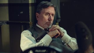 Tim Roth as Beckett sitting with his hands on his chest in Peaky Blinders: The Immortal Man. 
