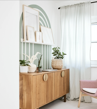 green room with a white rainbow decorative feature on the wall, wooden side table with plants on top, and a white floating shelf full of artwork, with a white/green linen curtain against the window
