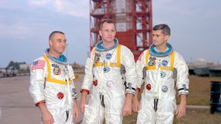 three men in white space suits stand outside in front of a launch pad tower