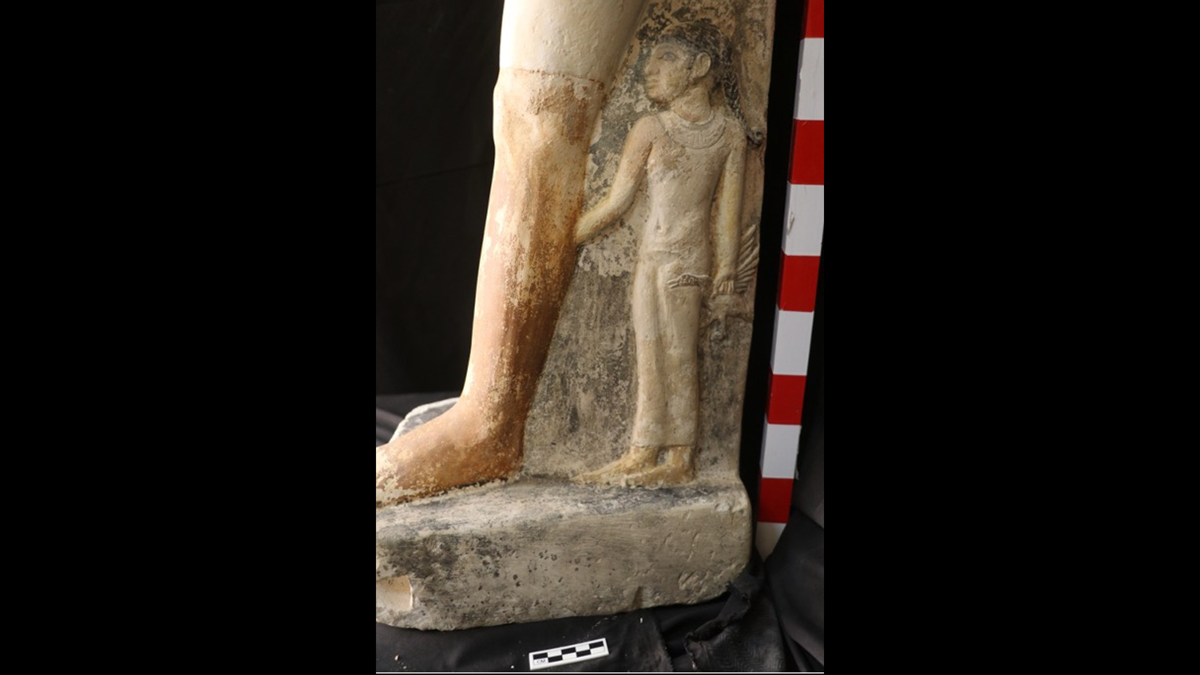 The side of a statue of a man. We only see his leg, which has a tiny girl holding onto it. The girl is holding a goose.