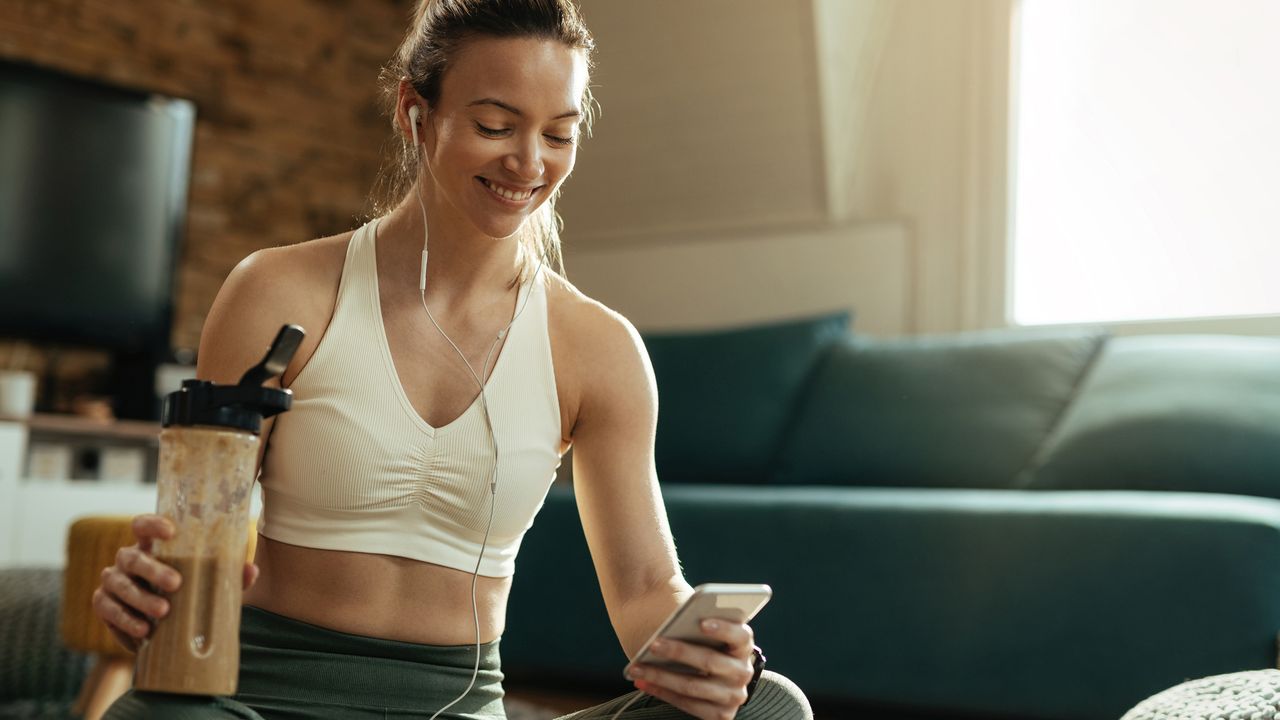 Happy athletic woman using mobile phone while drinking a protein shake at home